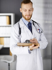 Friendly red-bearded doctor standing and writing with clipboard in clinic at his working place. Perfect medical service in hospital. Medicine, healthcare