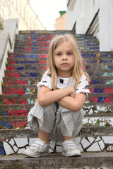 Stylish model girl six seven years old with blond hair sits on his haunches on a bright stone staircase on a city street in summer or spring.