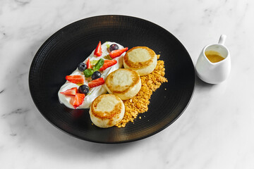 An ideal breakfast - Cottage cheese pancakes with white sauce, berries, strawberries and crunch in a black plate on a marble background. Ukrainian syrniki