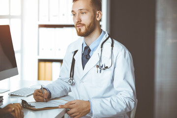 Friendly red-bearded doctor and patient woman discussing current health examination while sitting...