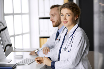 Friendly female doctor sitting in clinic with her male colleague at the background. Perfect medical service and medicine concept