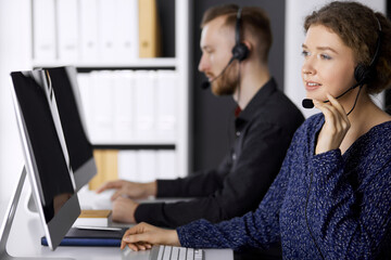 Businesswoman talking by headset while sitting with red-bearded colleague in modern office. Telemarketing and customer service. Diverse people group in call center