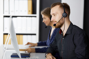 Bearded businessman talking by headset near his female colleague while sitting in modern office....