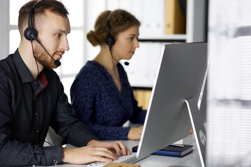 Fototapeta na wymiar Bearded businessman talking by headset near his female colleague while sitting in modern office. Diverse people group in call center. Telemarketing and customer service