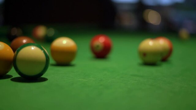 Kicking of colorful billiard balls with numbers with long wooden pool stick on table covered with green fabric in semi-dark room extreme closeup.
