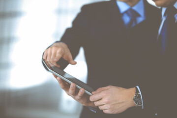 Business people use a tablet computer for discussion of their new project, standing in a modern office. Unknown businessman or male entrepreneur with a colleague at workplace. Teamwork and partnership