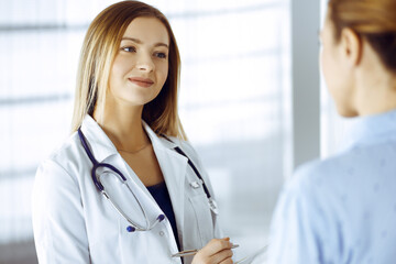 Young woman-doctor and her patient are discussing patient's current health examination, while standing together in a hospital office. Female physician is writing some marks, using a clipboard. Perfect