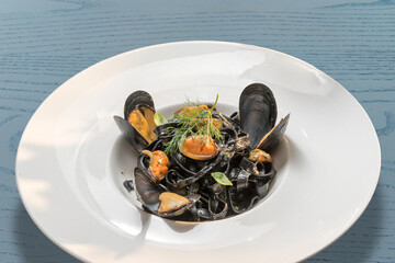 black pasta squid ink with mussels - italian tagliatelle al nero di seppia - in a white dish on blue wooden table