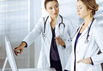 Two female physicians are discussing medical therapy, while standing at the table in a clinic office. Doctors use pc computer at work. Teamwork in medicine