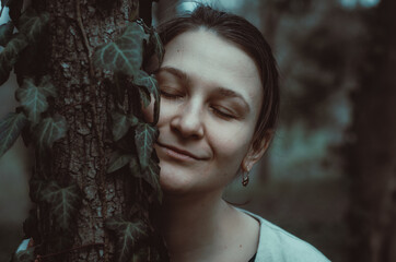 Moody atmospheric portrait of caucasian woman with eyes closed, smiling,  connecting with nature in a forest, face touching a tree. 	