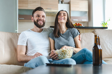 Young cute casually dressed couple in love having fun watching comedy movie on tv and eating pop...