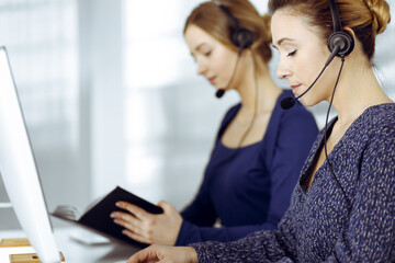 Two busineswomen have conversations with the clients by headsets, while sitting at the desk in a modern office. Diverse people group in a call center. Telemarketing and customer service
