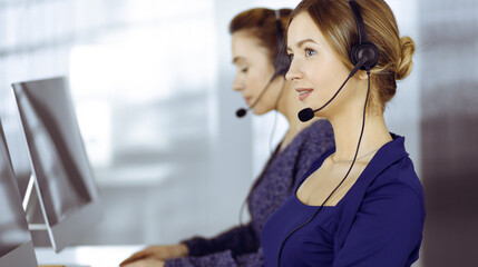 Obraz premium Two busineswomen have conversations with the clients by headsets, while sitting at the desk in a modern office. Diverse people group in a call center. Telemarketing and customer service