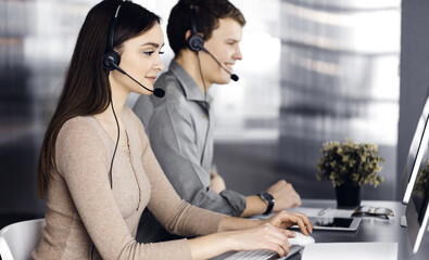 Two young people in headsets are talking to the clients, while sitting at the desk in an office. Focus on woman. Call center operators at work