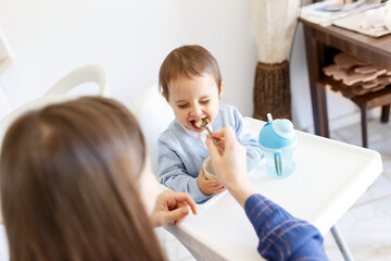 Mother feed little toddler baby boy happy and smiling with open mouth from plastic spoon in high chair view from back