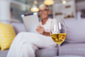 Happy retired senior woman relaxing at home, using digital tablet.Focus on wineglass