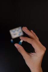 Hand holding a pill capsule close-up on a black background. The package is a blister with tablets in full focus.
