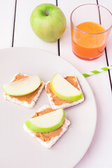 Healthy breakfast with peanut butter and apple sandwiches on rice cakes, carrot juice and apple on white wooden background