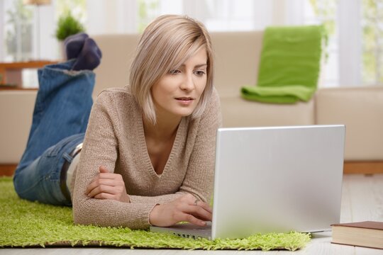 Young blonde woman lying on floor at home working on laptop computer.