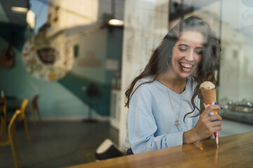 Chica joven tomando helado en cafeteria vista a través de un cristal
