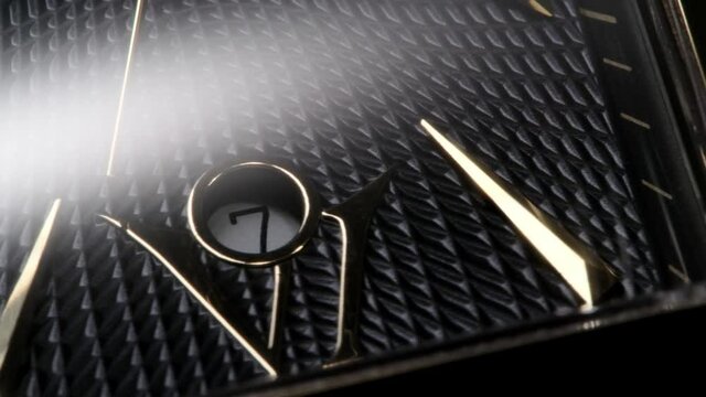 Luxury golden swiss made watch with square body and waffle pattern on clockface rotating while second arrow is passing by the date and numbers. Closeup with flashing light