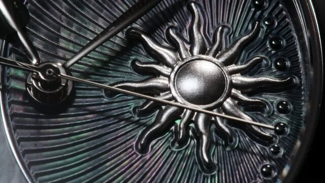 Silver second arrow running on nacrous ribbed clockface of rotating swiss made watch with sun. Extreme closeup of metallic details and ribbed pattern, front view