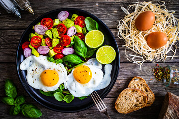 Sunny side up eggs with avocado, cherry tomatoes, toasted bread and red onion on wooden table

