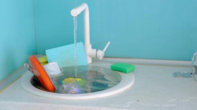 Full sink of dirty dishes in the kitchen. Open tap water runs from the sink to the countertop. The flood. 
