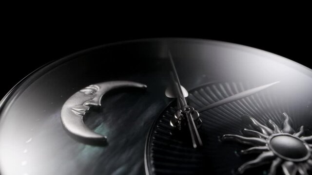 Silver second arrow running on nacrous clockface of rotating swiss made watch with moon and sun. Closeup of metallic details and ribbed pattern on black background