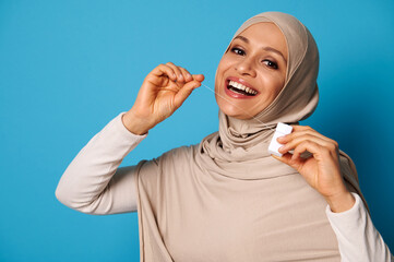 Cute Arab woman with covered head using a dental floss and posing over blue background
