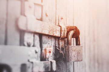 On the gray old wooden gate is a rusty latch, on which hangs an old metal lock, illuminated by sunlight. Closed.