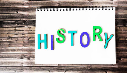 The word history is written in multicolored wooden letters on a white notebook. Wooden background.