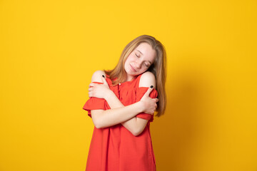 Cute teen girl hugs herself touches shoulders expresses self love smiles pleasantly dressed in summer red dress.