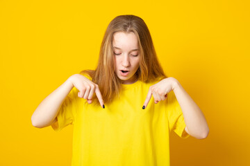 Amazed young girl points down with fore fingers, openes mouth from amazement, dressed in yellow...