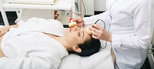 Beautiful brunette woman during facial microcurrent procedure for lifting skin with her beautician....