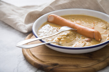 Deftige Deutsche Erbsensuppe als Eintopf mit Bockwurst in Emaille Teller auf Holzbrett und Marmor Hintergrund hell