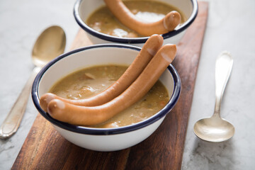 Zwei Emaille Schalen deftige Deutsche Erbsensuppe als Eintopf mit Wiener Würstchen auf Holzbrett und Marmor Hintergrund hell