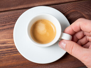 close-up of a man's hand holding a small white cup of aromatic espresso over a white saucer. Wooden background, Top view, flat lay