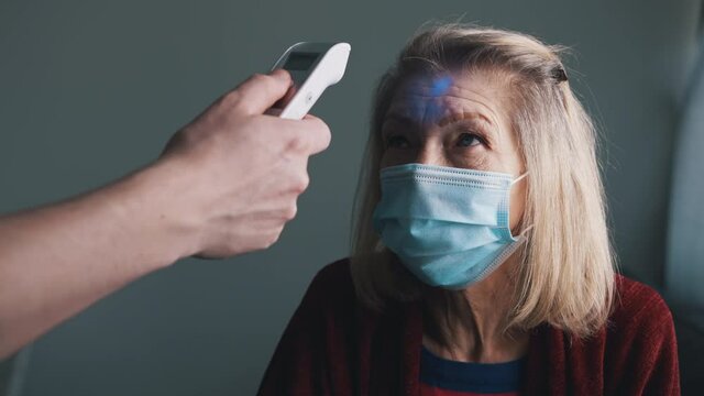 Check up of body temperature with digital thermometer. Old gray haired woman with face mask having dotor visit at home. High quality 4k footage