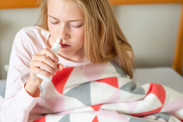 Sick teenage girl sitting on the bed covered with blanket using saline nasal spray