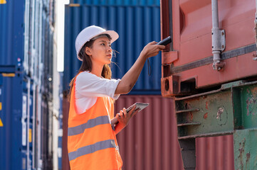 Smile Asian woman logistics worker wear safety helmet and protect suite working and checking product with taplets and scan code gun in shipping containerin commercial transport port