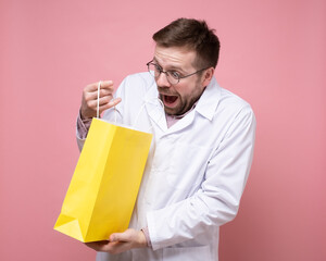 Young doctor received a gift, he holds a yellow package in hands, looks into it happily and is surprised. Pink background.