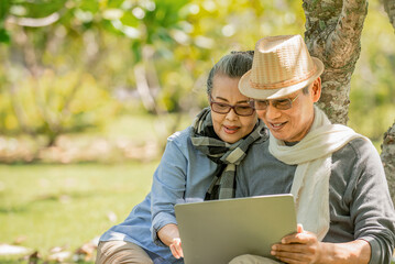 Senior asian couple  sitting under the tree  at park  using  laptop computer while holding coffee cup