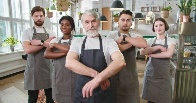 Portrait of mixed-race different male and female workers in aprons standing in cozy cafeteria looking at camera in good mood Restaurant business industry Senior cafe owner with young employees at work