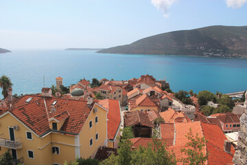 Naklejka na ściany i meble Top view of the orange tiled roofs of houses against the backdrop of azure sea water and beautiful mountains. Photography of Montenegro. Travel concept.