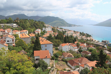 Naklejka na ściany i meble Top view of the orange tiled roofs of houses against the backdrop of azure sea water and beautiful mountains. Photography of Montenegro. Travel concept.