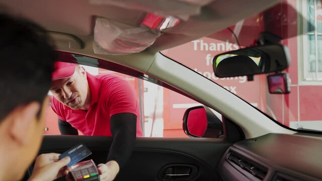 smiling gas station attendant collecting payment through window from young man at driver seat paying with credit card or qr code chatting with him.