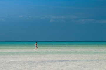 Fototapeta na wymiar A young woman walks on the water at low tide on Holbox Island in Mexico. In the background the cloudless blue sky. Concept travel adventures