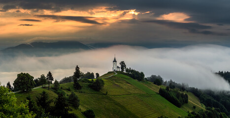Jamnik, Slovenia - Magical foggy summer morning at Jamnik St.Primoz hilltop church. at sunrise. The fog gently goes behind the small chapel with golden sky and Julian Alps at background - obrazy, fototapety, plakaty