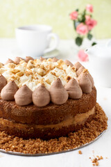 Closeup of a whole chocolate layer cake with caramel and nut topping on a light wooden background.